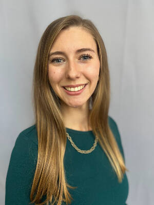 A person with long, straight, light brown hair wearing a green textured top and a gold chain necklace, standing in front of a neutral, light gray background.
